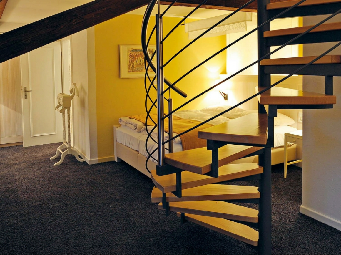 Ansicht eines Zimmer im Vordergrund ist eine Wendeltreppe mit offenen Holzstufen zu sehen, im Hintergrund steht ein Bett