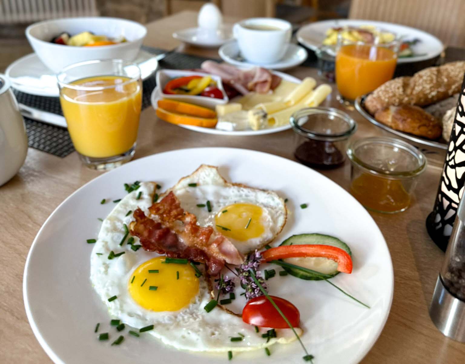 schön angerichteter Teller mit 2 Spiegeleiern im Vordergrund. Im Hintergrund gedeckter Frühstückstisch mit Orangensaft im Glas, Käseteller und weiteren Tellern und Tassen